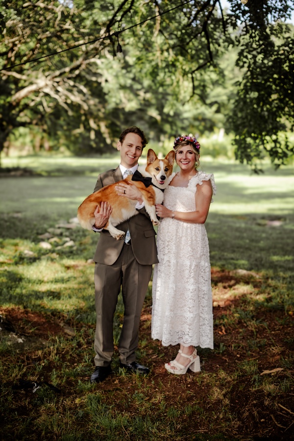 Justin, Leah, and Pip, on Leah and Justin's wedding day. Justin wears an olive suit, Leah wears a white lace dress, and Pip wears a black bowtie. All are smiling at the camera in a wooded, outdoor area.